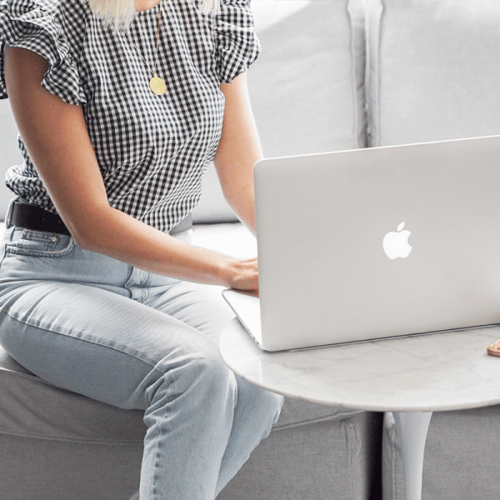 Person in gingham blouse and light colored jeans working on an Apple computer at a small table